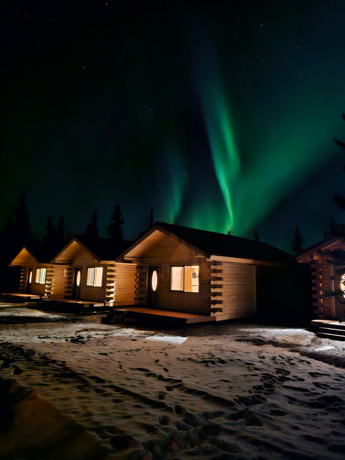 A row of log cabins is set against a night sky illuminated by vibrant green Northern Lights. Snow-covered ground reflects the ethereal glow, while the warm light from cabin windows offers a welcoming contrast to the surrounding darkness.