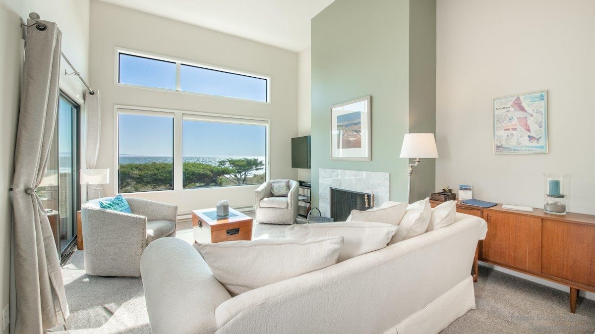 The living area features large windows allowing natural light to illuminate the space, showcasing views of the ocean and surrounding greenery. A cozy sofa and armchairs are arranged around a wooden coffee table, with a modern lamp providing additional lighting and a fireplace adding warmth.