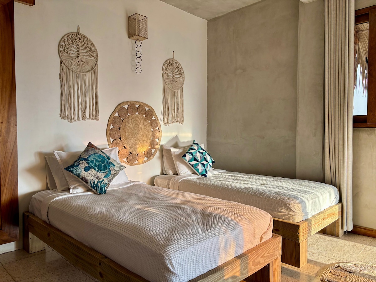 A well-arranged sleeping area is presented with two spacious beds, each featuring decorative pillows. Textured wall decor adds a subtle artistic touch, while natural light enters through a nearby window covered with light curtains.
