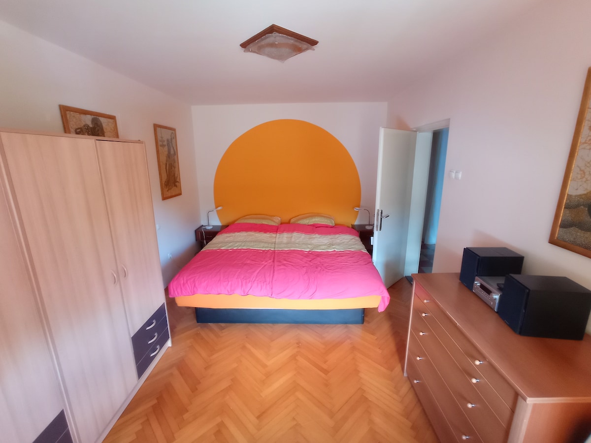 A bright bedroom features a large waterbed covered in vibrant pink and yellow bedding. The walls are complemented by a bold orange accent wall. Storage is available in a wood wardrobe and dresser, while bedside lamps provide functional lighting. A door leads to the shared hallway.