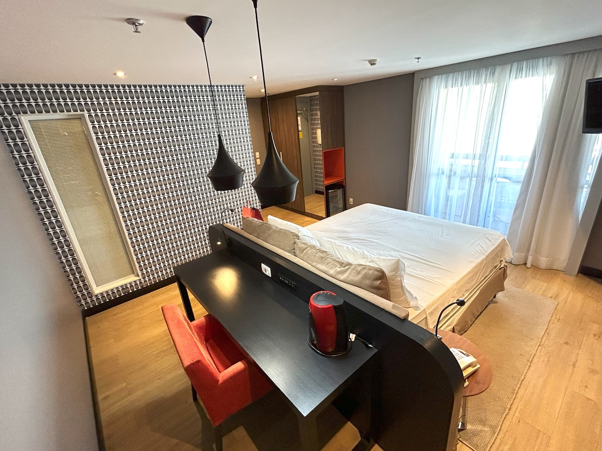 A spacious flat interior features a Queen bed with a minimalist design. A work desk with a comfortable chair and a red coffee maker are positioned nearby. Large windows allow natural light to fill the room, illuminating the modern decor and warm wooden flooring.