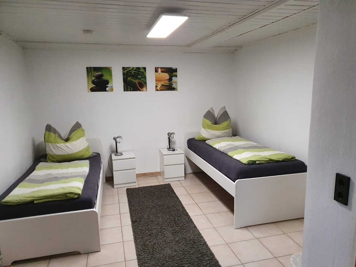 The sleeping area features two twin beds with gray linens, accented by green striped pillows. Each bed is paired with a white nightstand and a silver bedside lamp. The light-colored walls are adorned with three framed images, contributing to a calming environment.