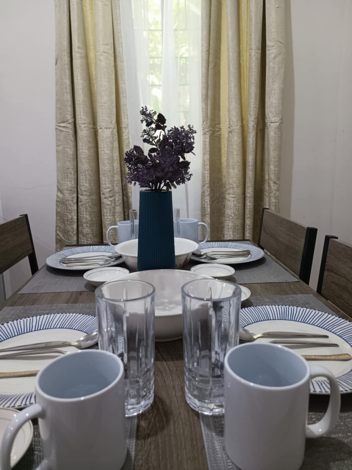 A dining table is set for four, featuring neatly arranged plates and cutlery. A slender blue vase holds fresh purple flowers at the center. Clear glasses and mugs are also placed on the table, with soft light filtering through sheer curtains.
