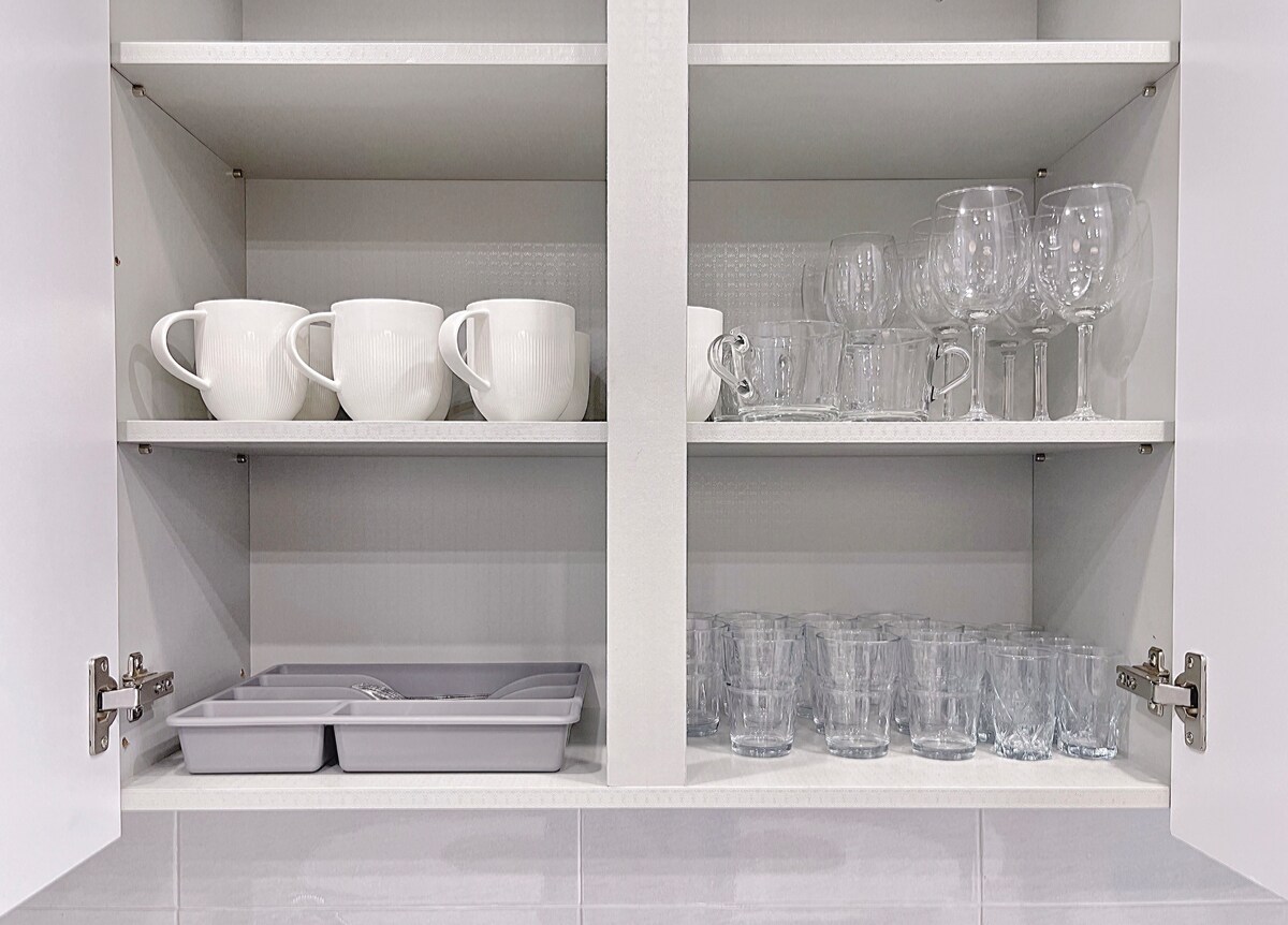 A kitchen cabinet is displayed with two levels of organized dishware. The top shelf contains white mugs and clear wine glasses, while the bottom shelf features additional glassware, including tumblers, arranged neatly. A tray for utensils is positioned on the bottom shelf.