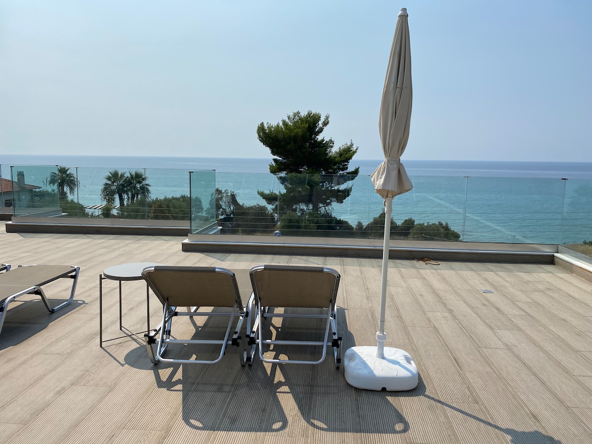 Two lounge chairs are positioned on a spacious deck, facing the sea. A small table is placed nearby, accompanied by a large umbrella on a stand. The expansive ocean view is visible in the background, with trees framing the area.
