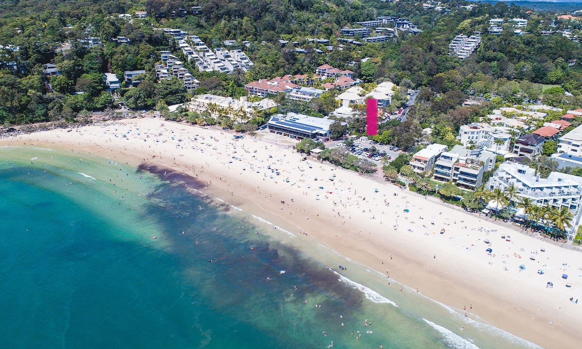An aerial view captures Noosa's Main Beach, featuring sunlit sandy shores and gentle waves. Sunbathers and beachgoers populate the beach area, while lush greenery lines the background, contrasting with buildings and amenities nearby.