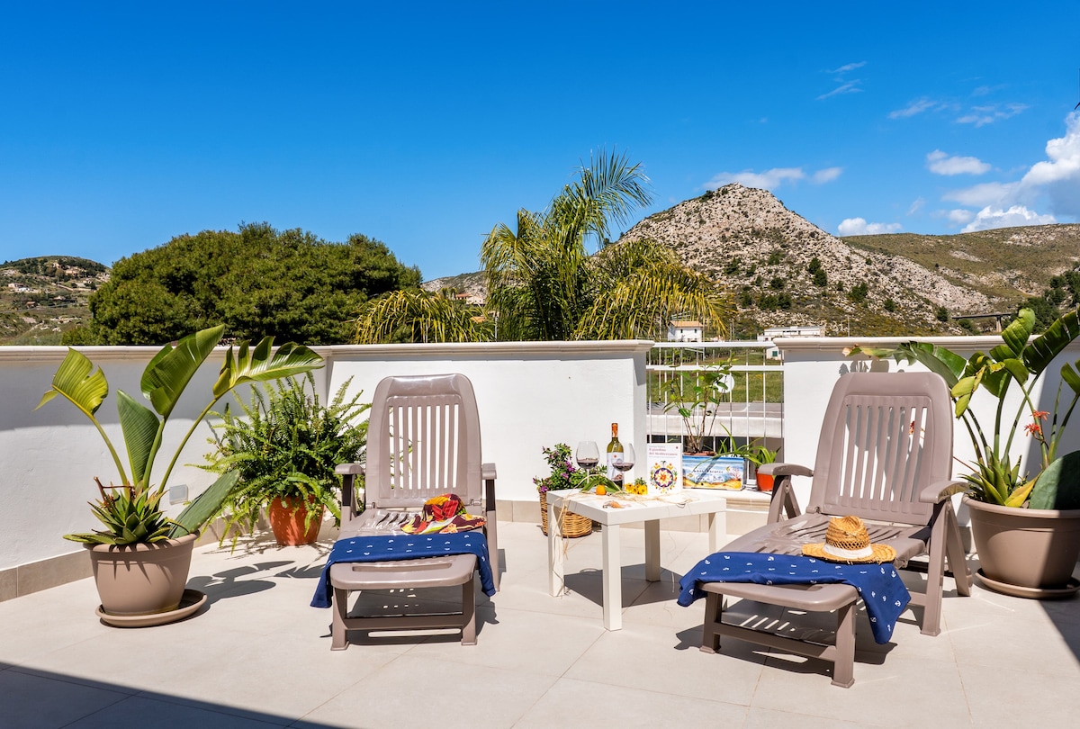 A terrace is presented, featuring two lounge chairs with light covers, a small table, and a selection of plants. The backdrop showcases rolling hills and a clear blue sky, creating a serene outdoor setting. Bottles and decorative items are arranged on the table.