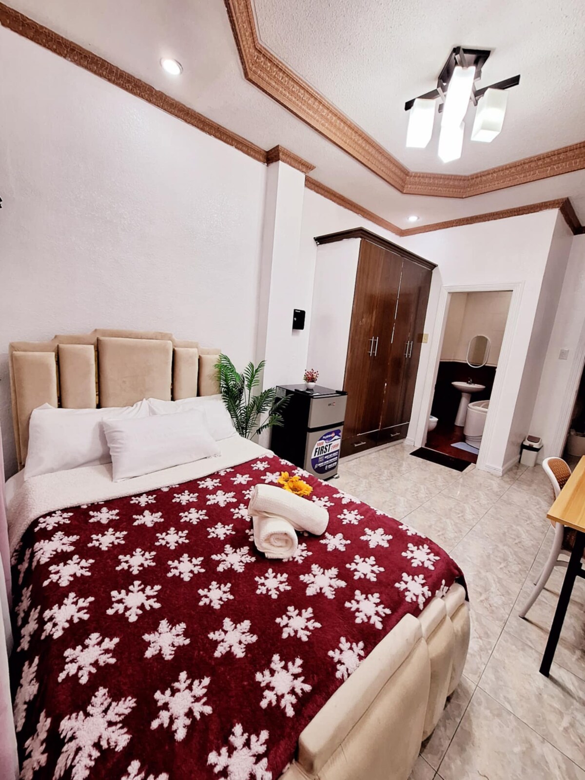 A cozy sleeping area features a spacious bed covered with a patterned blanket. A plant is placed near the bed, and a refrigerator is visible. A dark wooden wardrobe and a dining table are present. A bathroom door can be seen in the background.