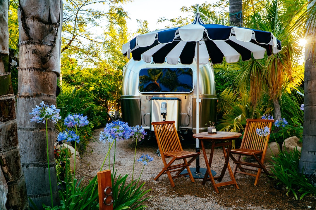 A cozy outdoor seating area features two wooden chairs beside a round table, surrounded by vibrant purple flowers and lush greenery. An Airstream is positioned under a black and white striped umbrella, adding a charming touch to the tranquil garden setting.