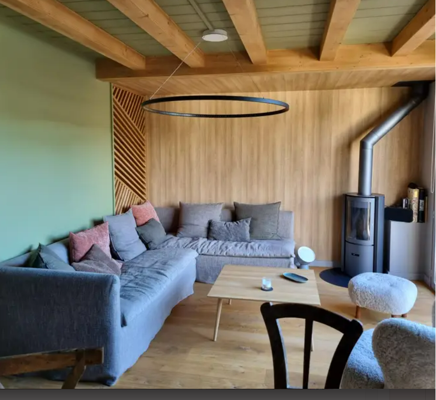 A cozy living area features a large sectional sofa adorned with various cushions, complemented by a light wooden table. A wood stove stands in the corner, while a distinctive circular light fixture hangs from the wooden beam ceiling, adding a touch of elegance.
