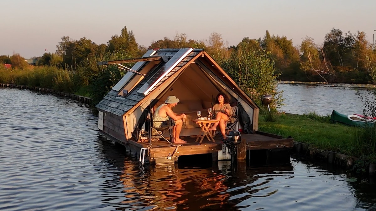A wooden floating cabin is situated on calm waters, surrounded by greenery. The structure features an angled roof with solar panels and a deck area with two chairs and a small table. Canoes are visible nearby, suggesting opportunities for exploration.