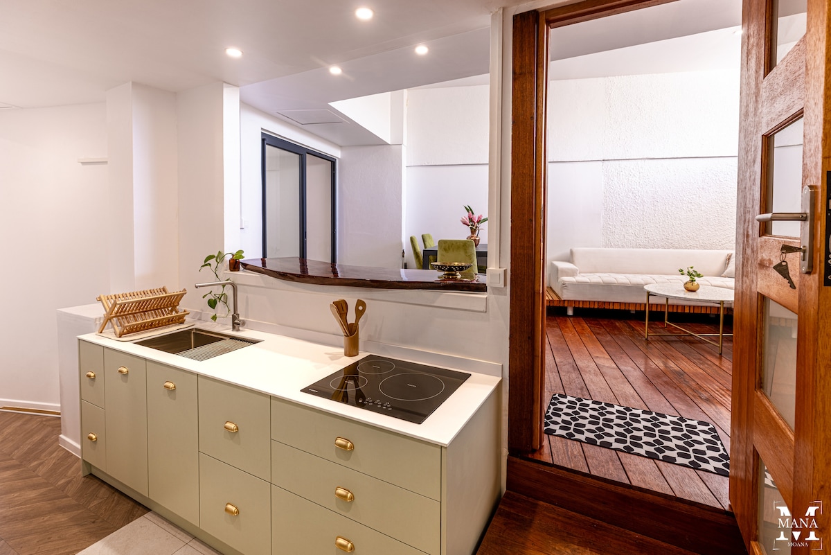 The kitchen features a modern layout with a smooth countertop and built-in cooking appliances. Soft green cabinetry provides storage, while a wooden drying rack adds functionality. A doorway leads to a bright seating area, adorned with natural light and comfortable furnishings.