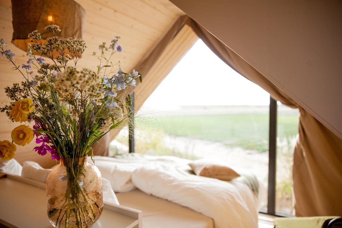 A vase filled with wildflowers is placed on a bedside table, with a spacious bed visible in the background. Natural light filters through a large window, revealing green fields outside. The interior features wooden walls and a soft color palette, creating a serene environment.
