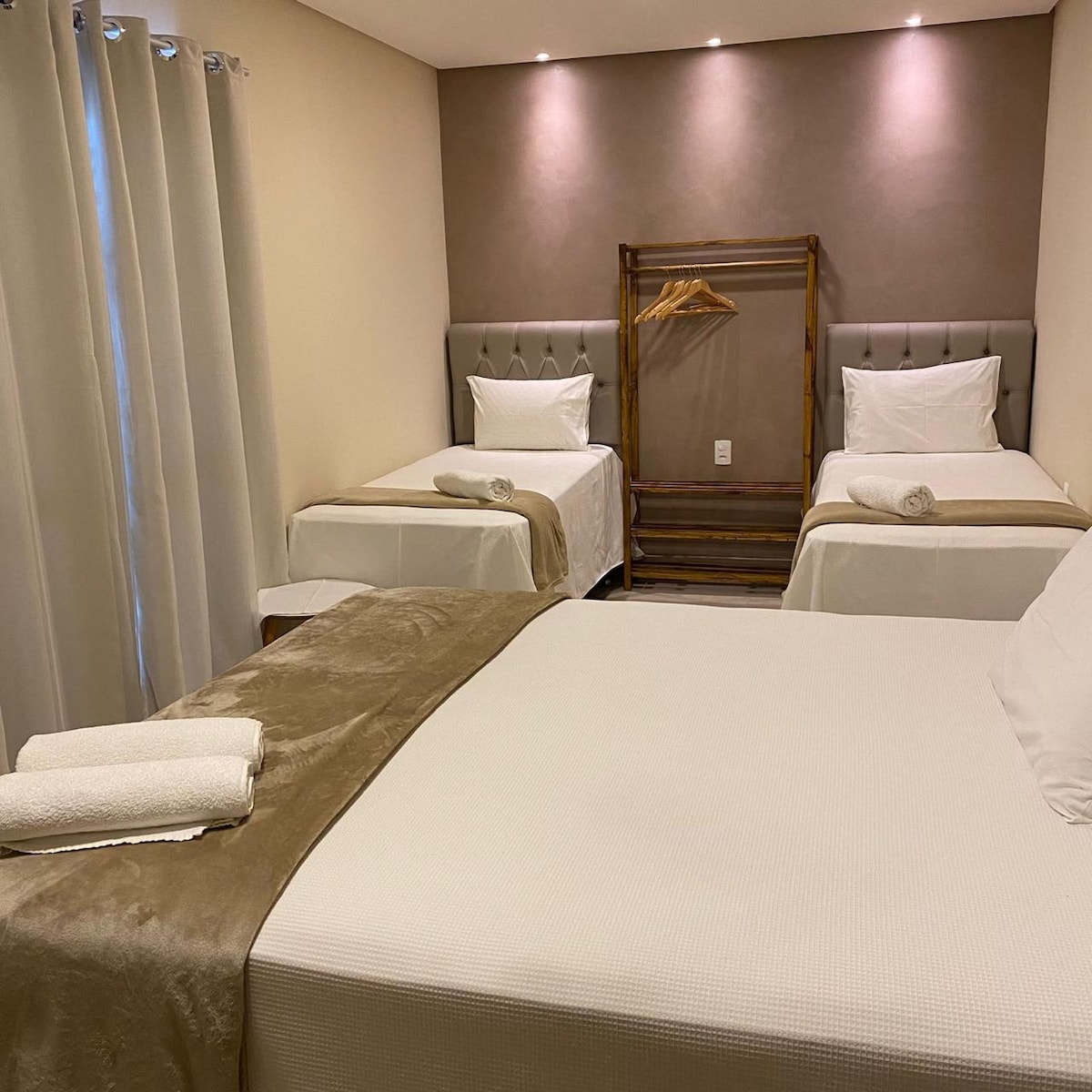 The room features three beds arranged with neutral-colored linens and soft blankets. A clothing rack with wooden hangers is positioned against a textured wall. Natural light enters through a partially draped window, and soft lighting highlights the serene atmosphere.