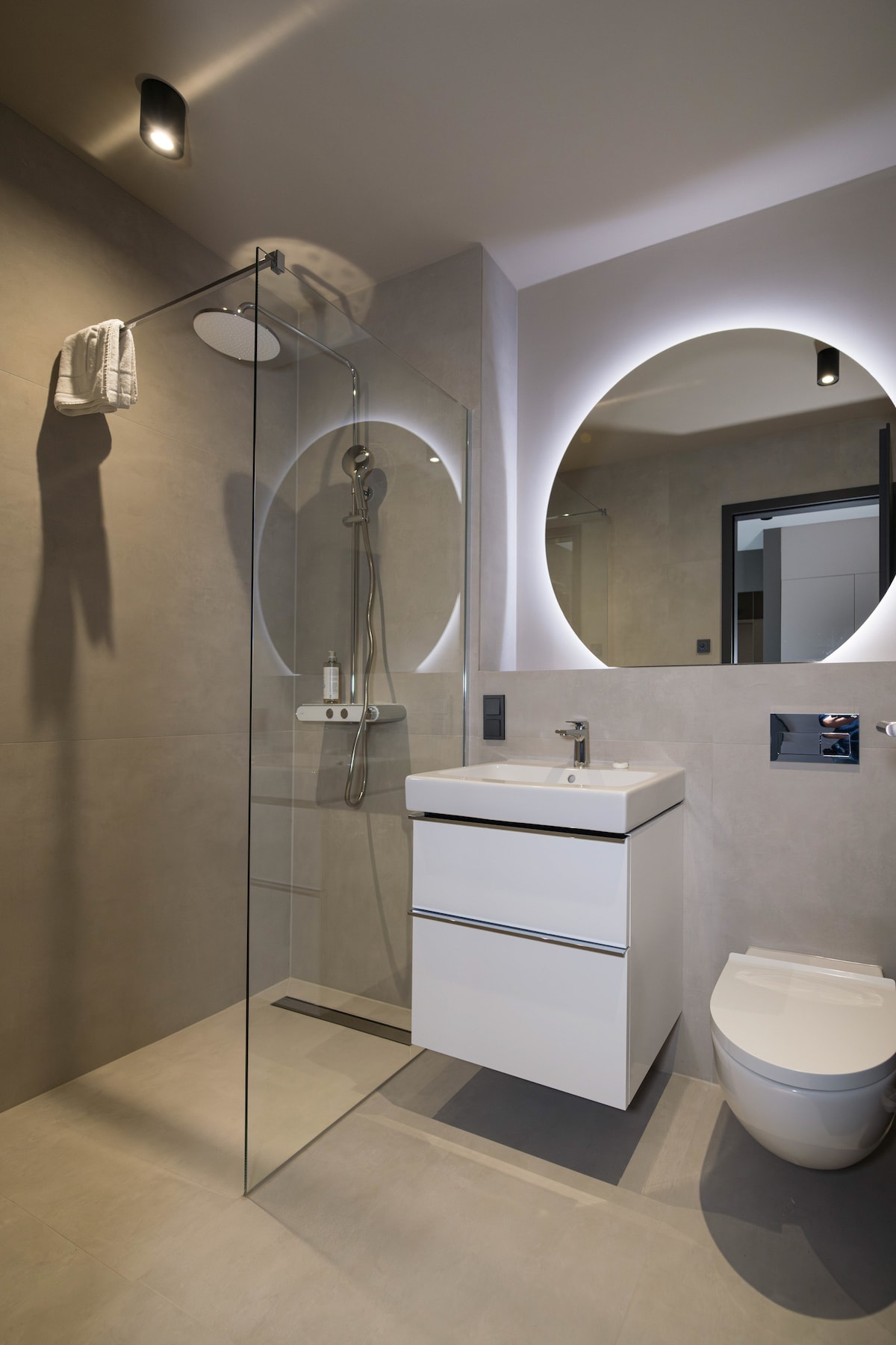 The modern bathroom features a glass shower with a rain showerhead and minimalist design elements. A sleek white vanity with storage sits below a round mirror, and a toilet is positioned discreetly. Soft lighting enhances the neutral-toned surfaces throughout the space.