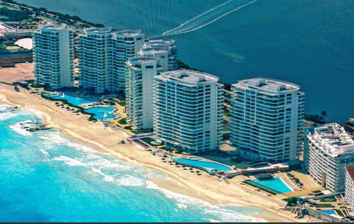 An aerial view captures a series of elegant beachfront condominiums, with expansive pools nestled along the shore. The blue Caribbean Sea contrasts with the golden sandy beach, while green coastal vegetation is visible in the background, creating a serene seaside atmosphere.