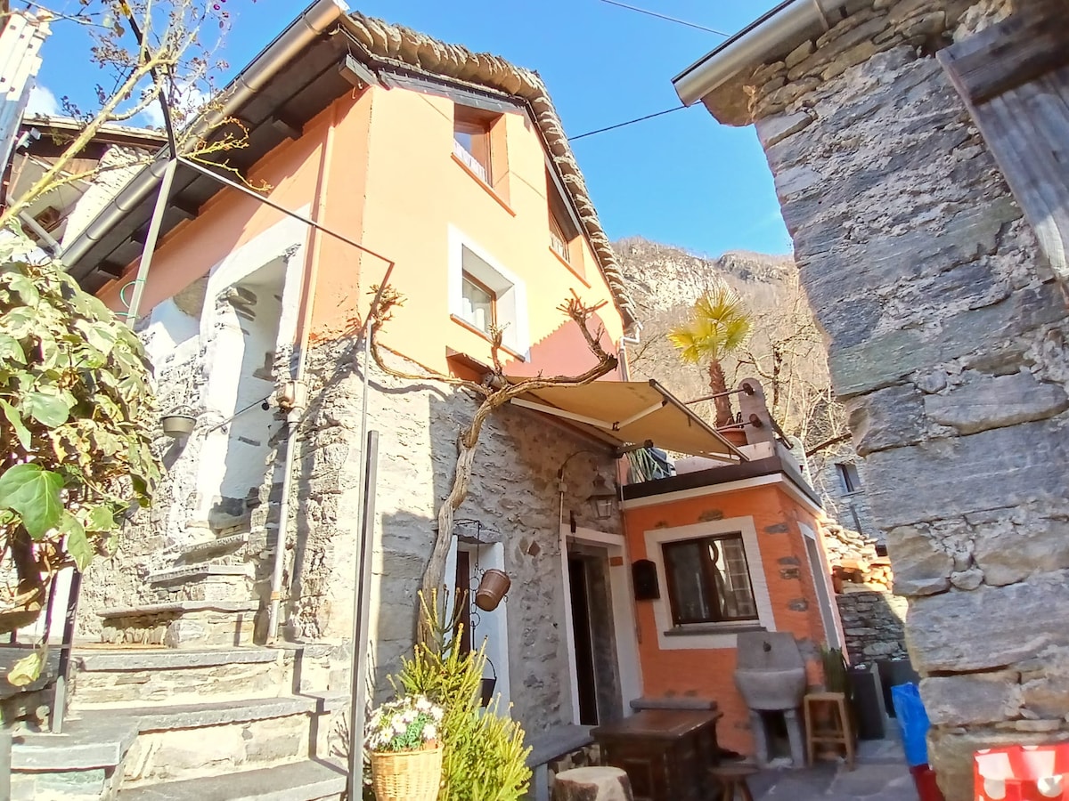 A rustic three-story house is depicted, with warm earth-toned walls and a sloped roof. Steps lead up to the entrance, flanked by greenery. A small outdoor seating area with wooden furniture is visible, providing a cozy space for relaxation. The surrounding stone buildings and mountainous backdrop enhance the setting.