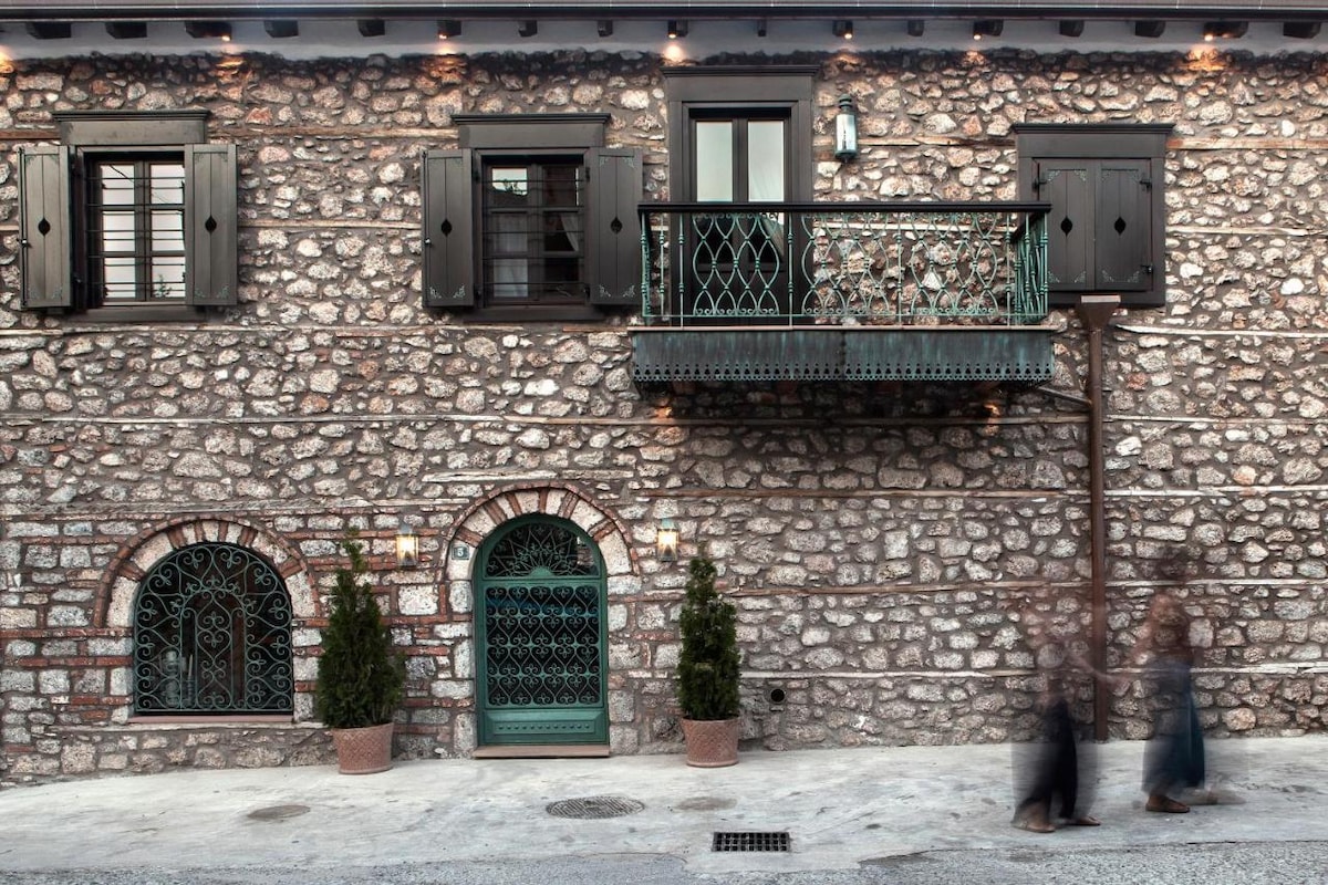 The historic stone facade is accentuated by dark wooden shutters and an elegant balcony. Decorative wrought iron elements can be seen framing two arched windows, while potted greenery adds a touch of nature to the entrance.