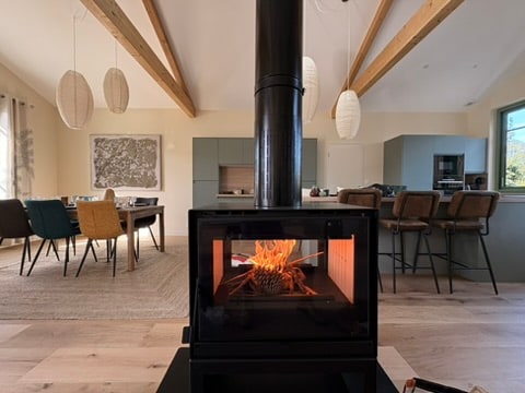 A modern living space showcases a stylish wood-burning stove as the focal point. Exposed wooden beams frame the space, which features a dining area with a mix of colored chairs and a kitchen in the background. Natural light enters through a nearby window.