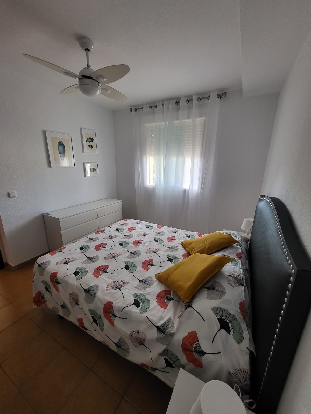 A bright sleeping area features a spacious bed adorned with a colorful floral bedspread and two yellow pillows. Light filters through sheer curtains, illuminating the room. A dresser is positioned against one wall, complemented by framed artwork hung above. A ceiling fan adds functionality to the space.