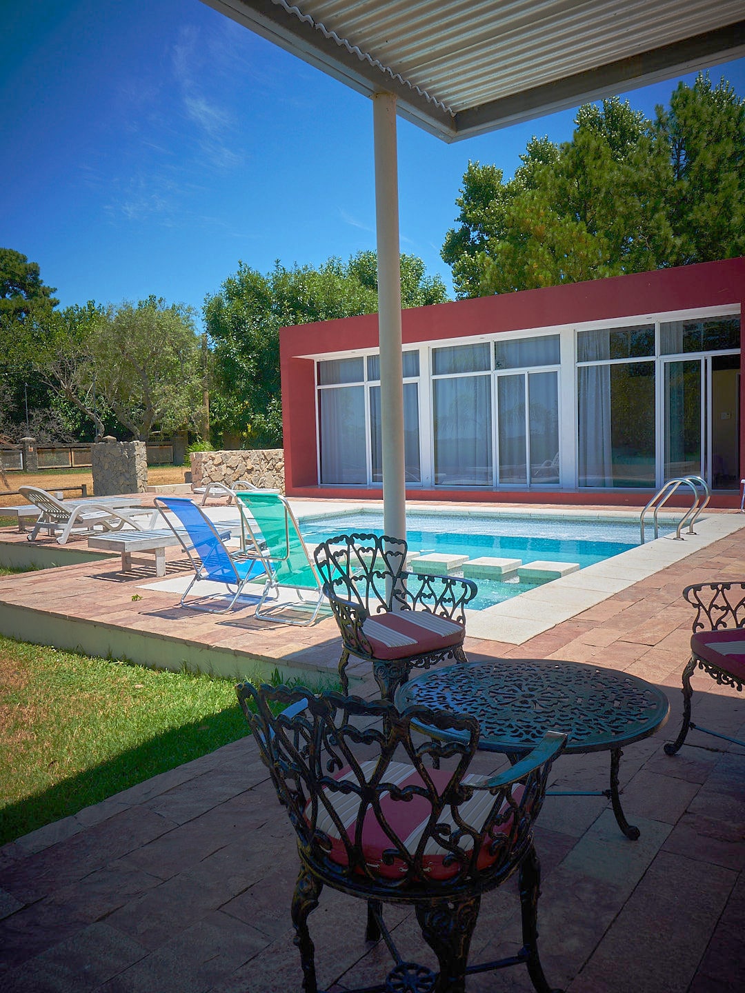 A serene outdoor space is presented, featuring a crystal clear pool surrounded by lounge chairs. A dining set with intricately designed chairs is positioned nearby. The backdrop includes large glass windows that reveal a spacious interior, with lush greenery enhancing the tranquil setting.