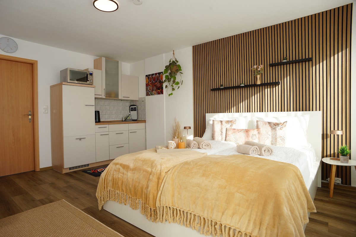 A bright and cozy room features a spacious bed, covered with a soft beige blanket. The wall behind the bed is accented with wooden panels. A small kitchenette is visible, equipped with essential appliances. Decorative plants and subtle artwork add a touch of warmth to the space.