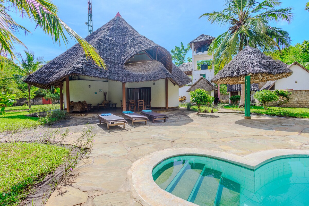 A traditional Swahili-African style building with a thatched roof is shown, surrounded by lush gardens. A private pool in a unique shape sits beside the stone patio, where sun loungers are arranged. The image captures a serene outdoor atmosphere under clear blue skies.