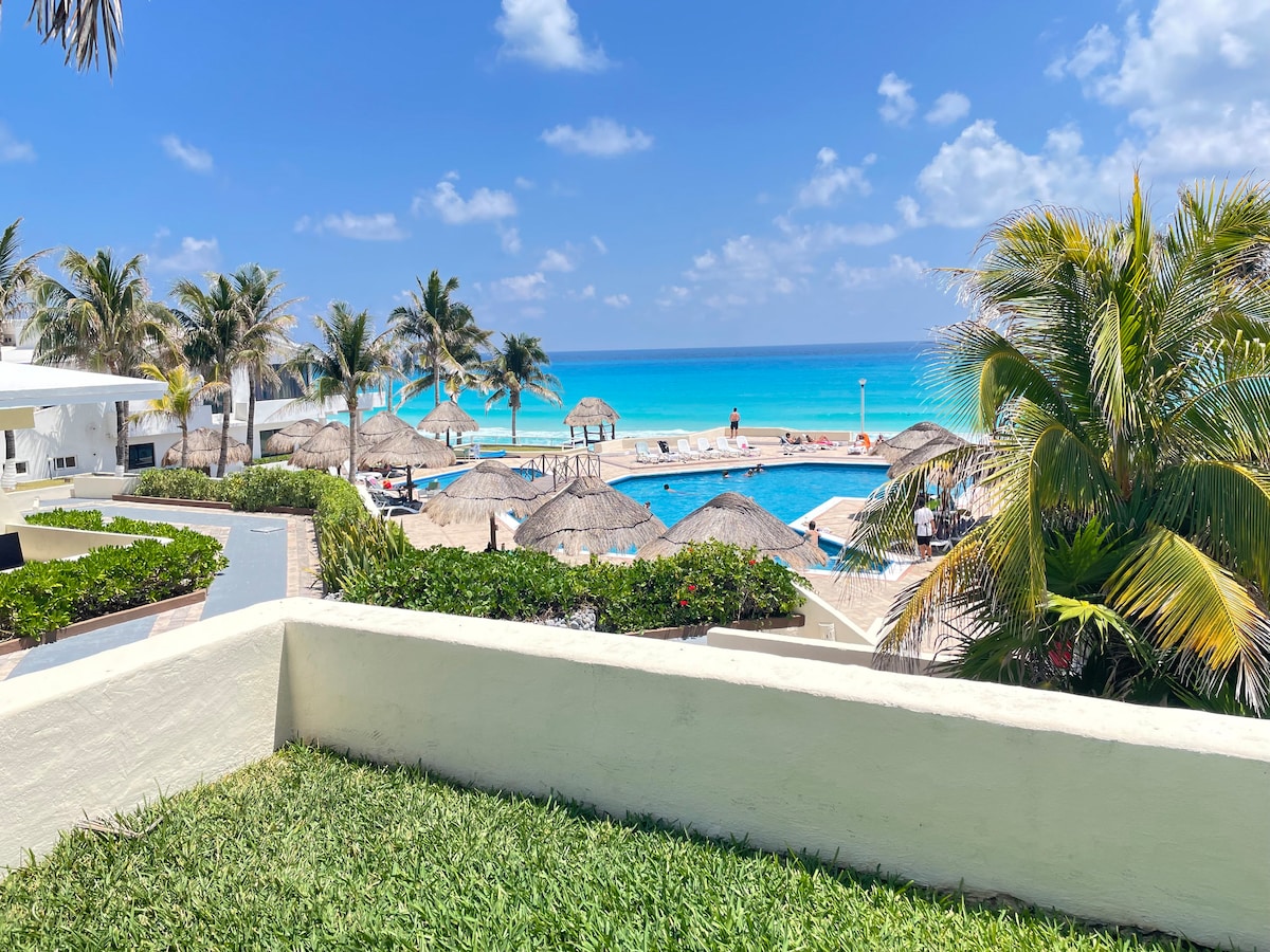 A view of the beach and swimming pool area, framed by lush green grass and tropical palm trees. Lounge chairs and palapas are set up along the sandy shore, with waves gently lapping at the shoreline under a clear blue sky.