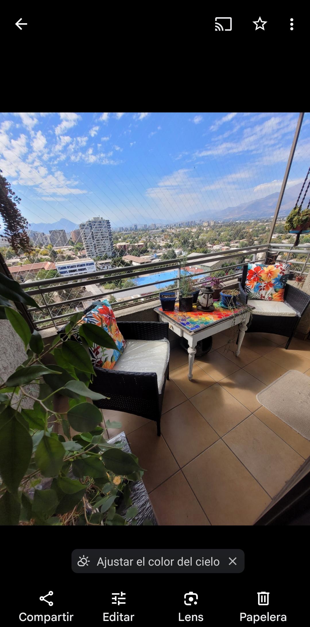 A balcony area is presented, featuring two comfortable chairs adorned with colorful cushions. A small table sits between them, accompanied by various plants. The view overlooks the city skyline and distant mountains under a partly cloudy sky.