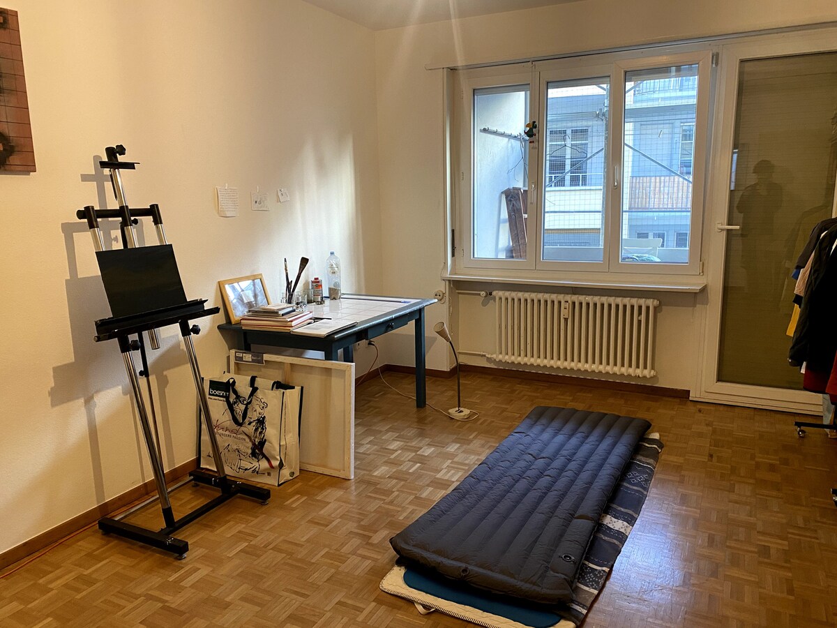 A single room features a work desk with an easel, accompanied by art supplies and a water bottle. A folding mattress is positioned on the wooden floor, and large windows allow natural light to brighten the space. A radiator lines the wall beneath the windows.