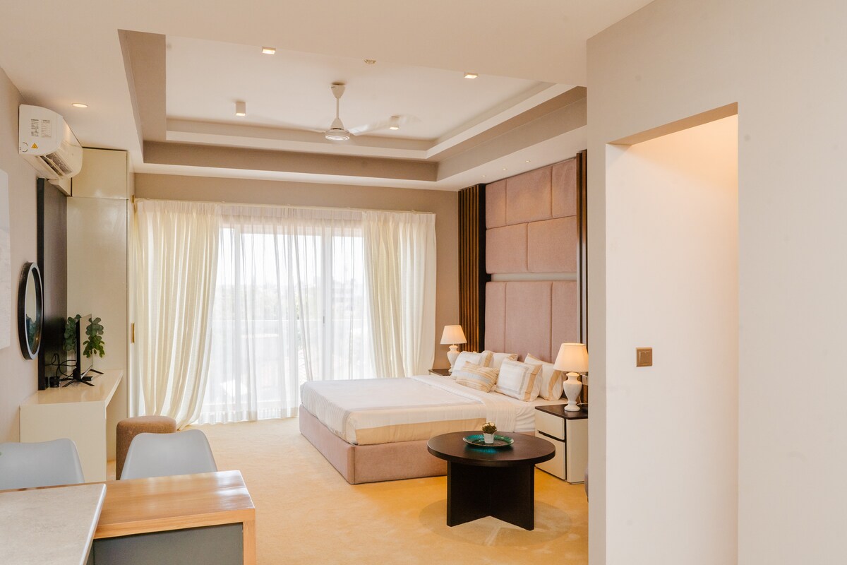 An open studio layout features a spacious king bed with fresh linens. Large windows allow natural light to fill the space, complemented by soft, neutral tones. A modern seating area includes a round table and chairs, while a TV is mounted on the wall.