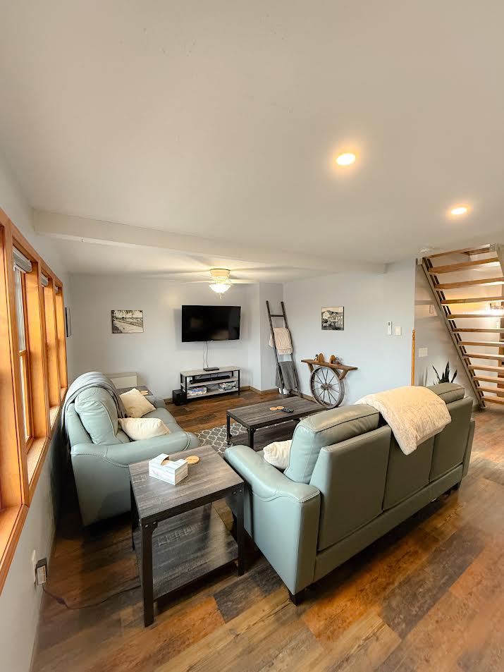 A comfortable living area is furnished with two light gray sofas and a dark wood coffee table. A television is mounted on the wall across from the seating. Natural light enters through multiple windows, and a stylish staircase is visible in the background.