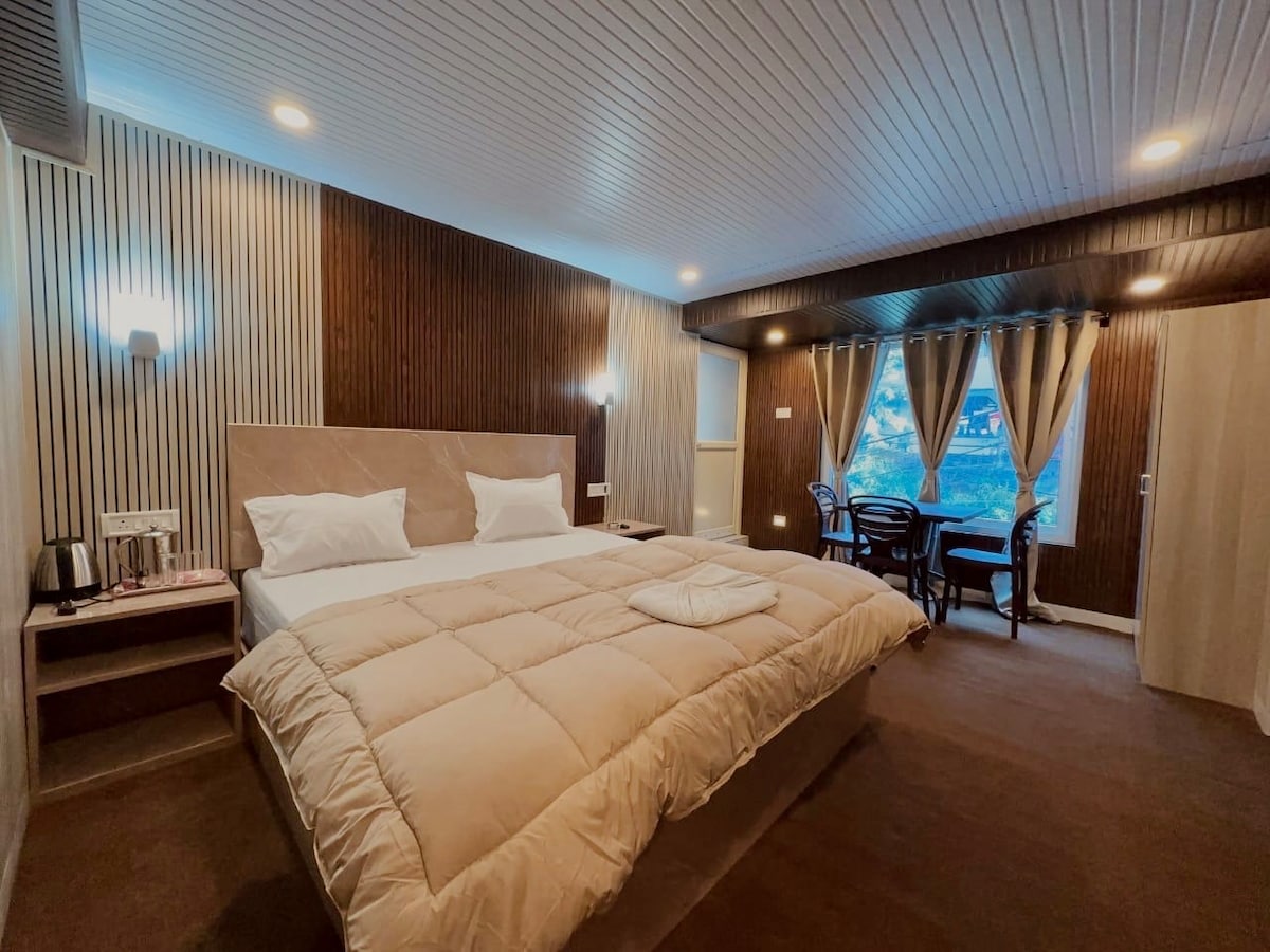 A spacious room features a large bed covered with a soft quilt, flanked by two bedside lamps. In the corner, a small table and chairs are positioned near a window dressed with sheer curtains, allowing soft light to enter the space.