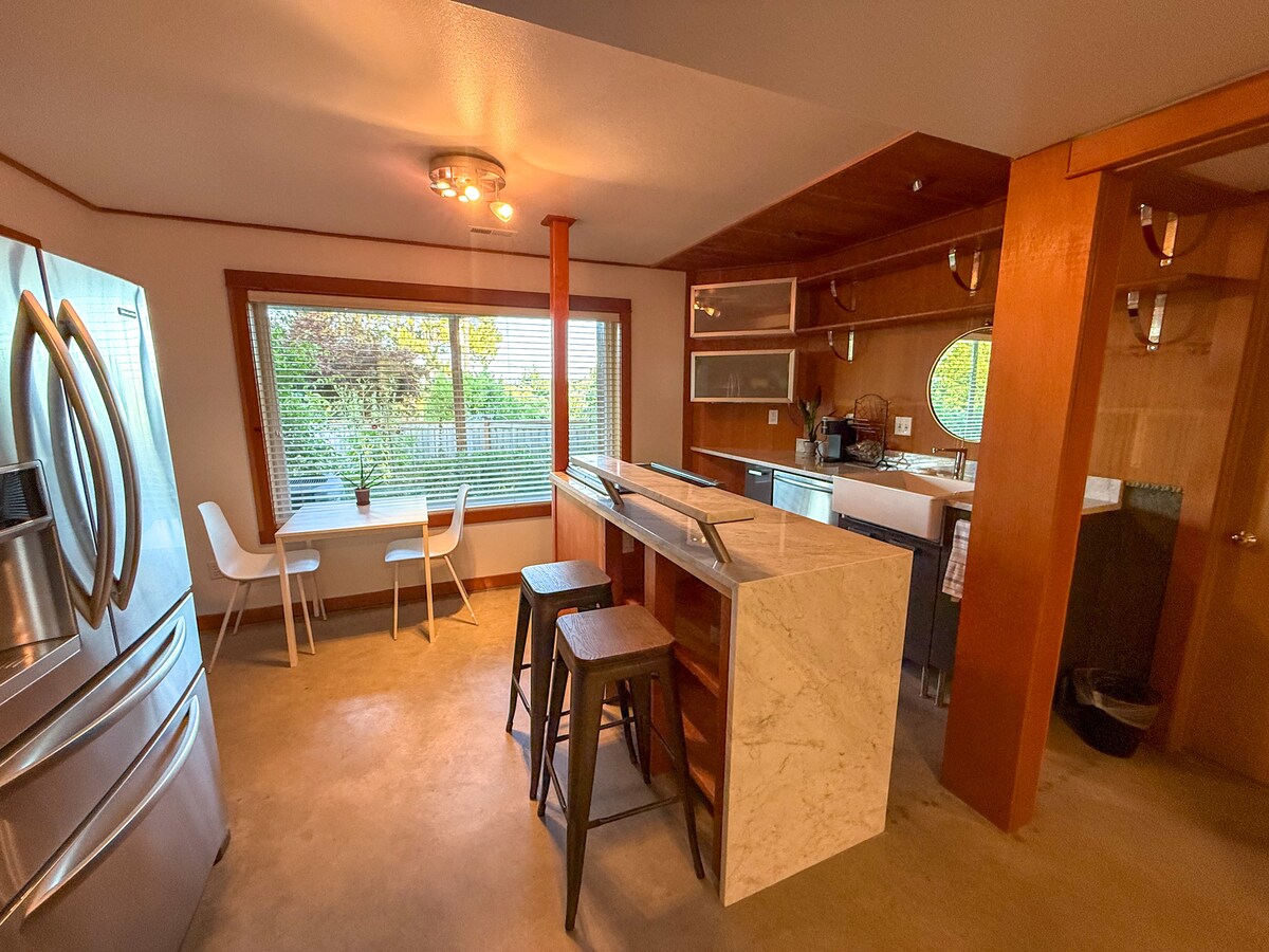 A modern kitchenette features stainless steel appliances, including a full-sized fridge and a small dining area with two white chairs and a table. Natural light filters through the nearby window, highlighting the wood accents and spacious countertops.
