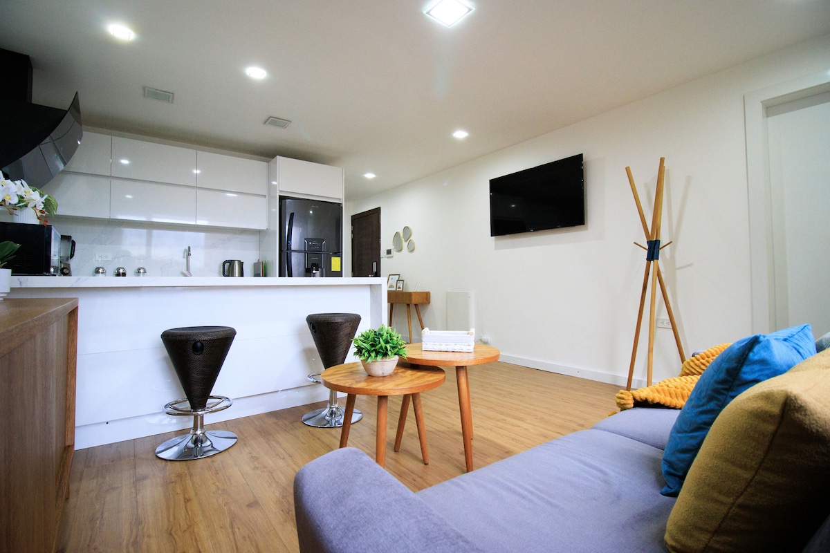 The living area features a strategically arranged sofa with colorful cushions, accompanied by a sleek coffee table. A modern kitchenette is visible in the background, showcasing white cabinetry and stainless-steel appliances. A flat-screen TV is mounted on the wall, enhancing the functionality of the space.