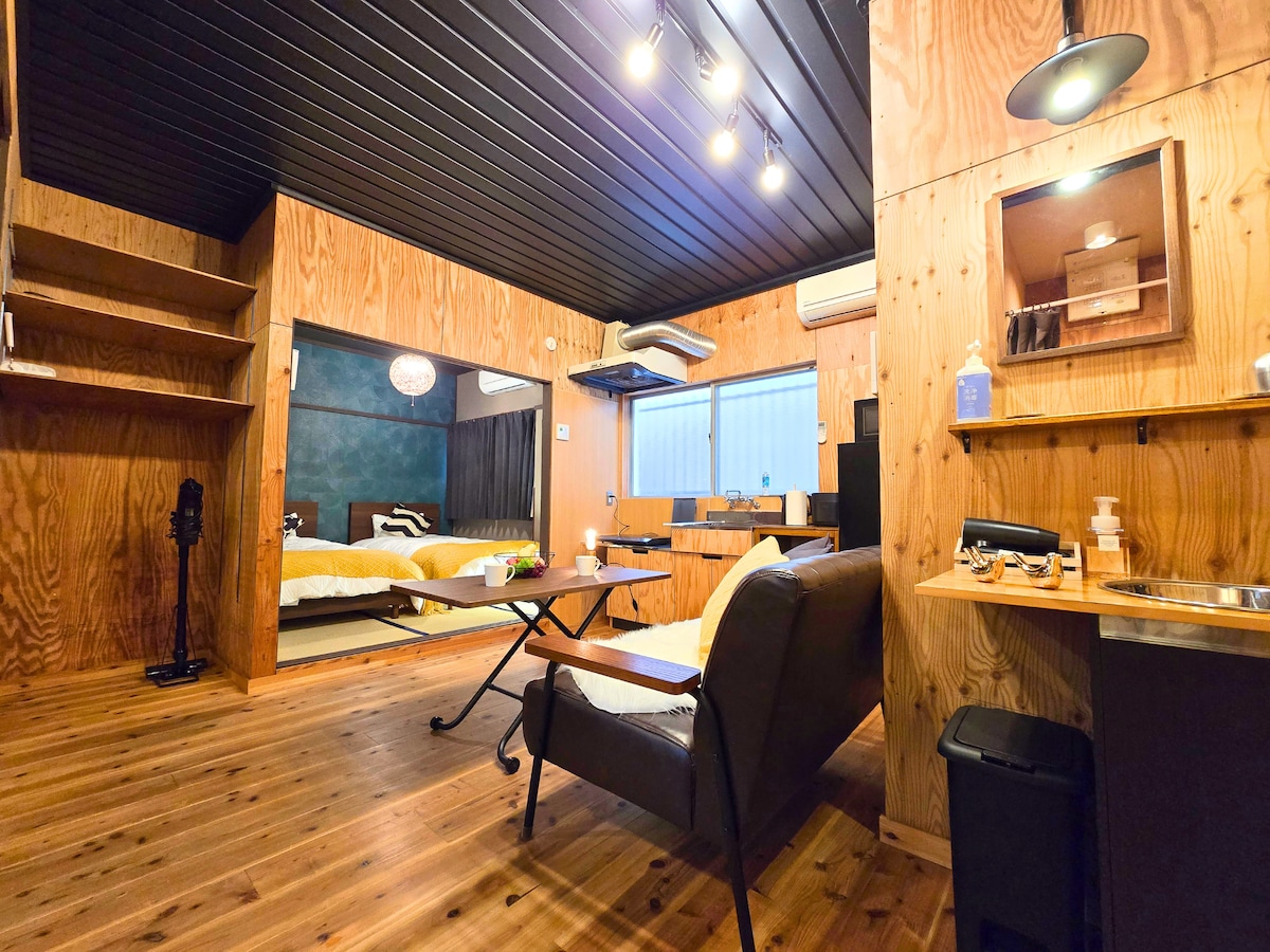 A spacious interior is depicted, featuring wooden walls and flooring. Two single beds are visible in the background, separated by a light curtain. In the foreground, a seating area includes a comfortable chair and a small table. The kitchen area is equipped with counter space and essential appliances.
