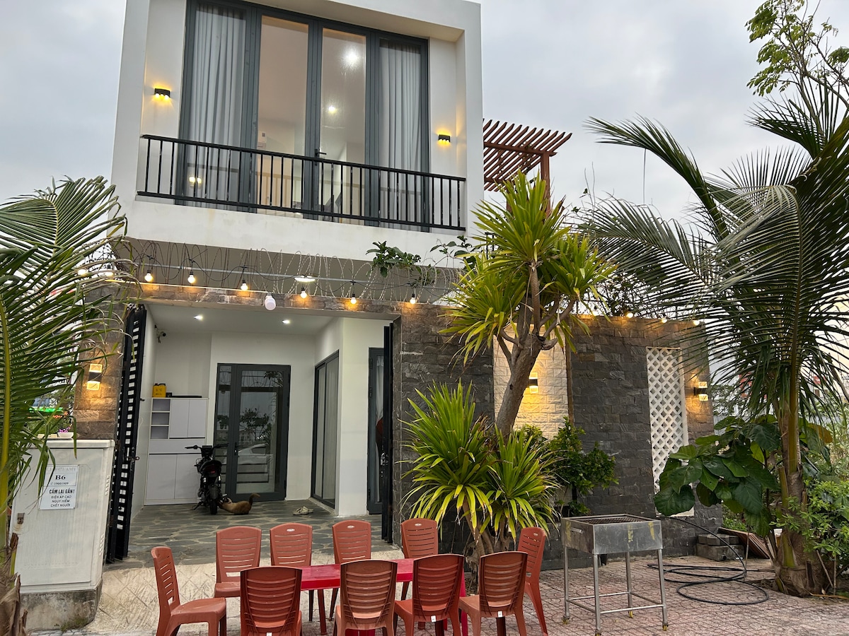 The exterior of a modern two-story house is shown, with expansive glass doors and a balcony above. Outdoor seating is arranged around a table, surrounded by landscaped greenery and decorative lights. A stone wall complements the design, creating an inviting outdoor area.