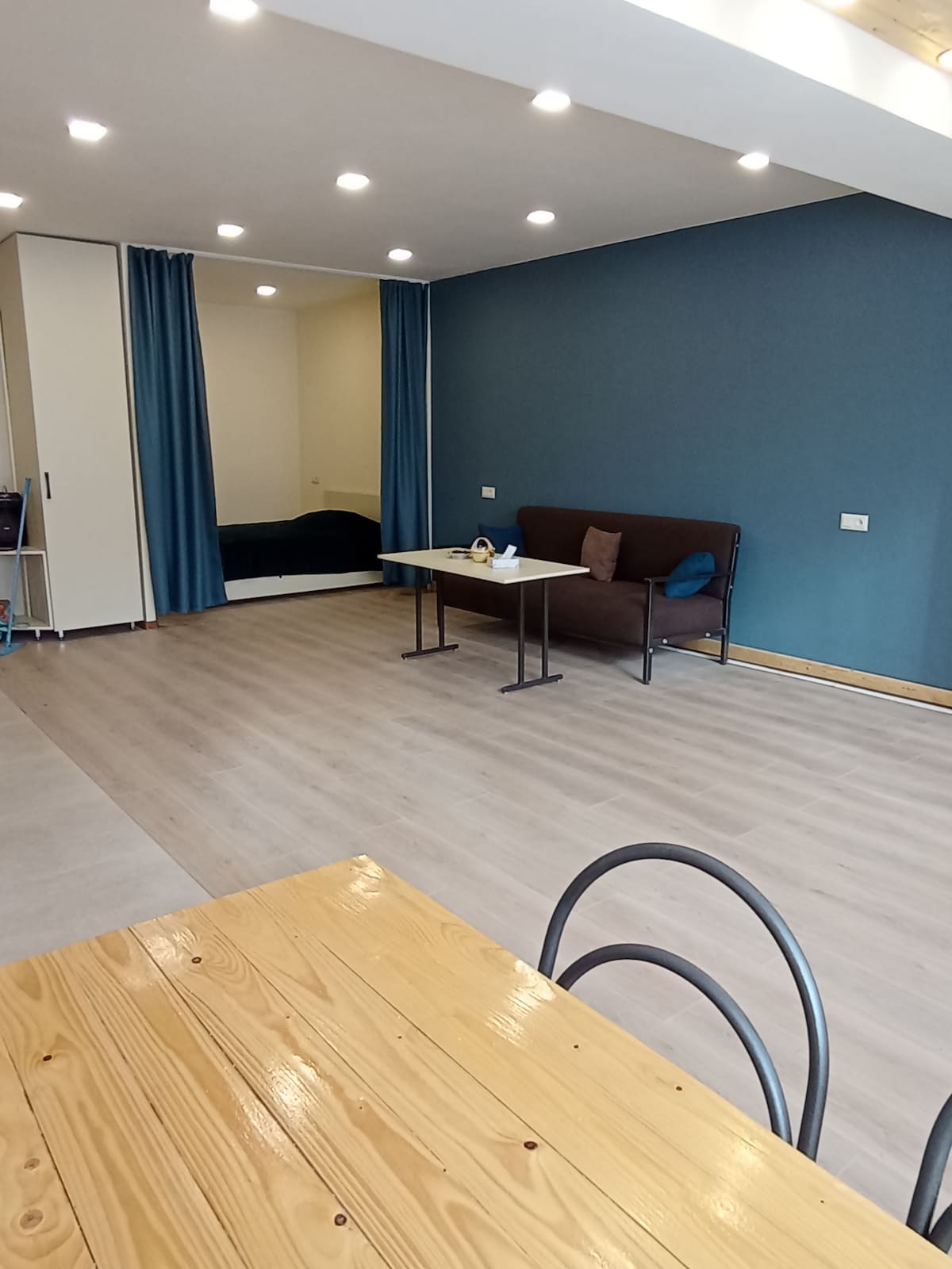 An open living space features a wooden dining table paired with black chairs, complemented by a brown sofa. A large mirror reflects a nearby sleeping area, while blue curtains add a touch of color against the light blue wall. Ample lighting ensures a bright atmosphere.