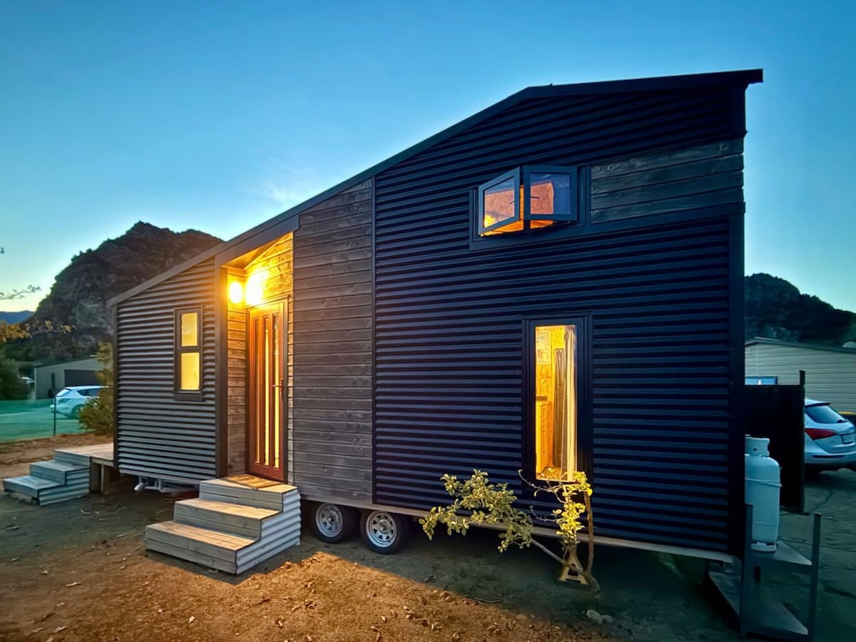 The exterior of the tiny home is shown at dusk, with warm light emanating from the porch and windows. The structure features a modern design with a combination of dark siding and natural wood elements, complemented by steps leading to the entrance.