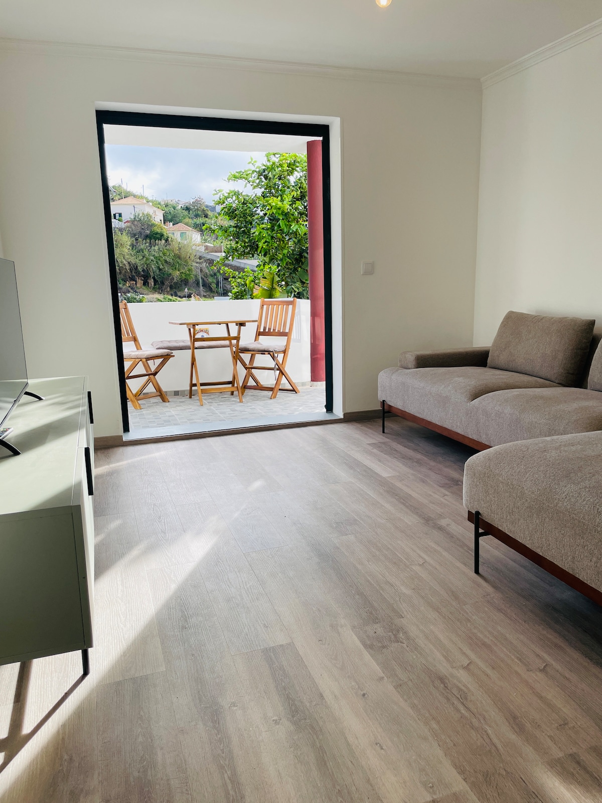 A well-lit living space features a cozy sofa and a television, with light wood flooring enhancing the atmosphere. A doorway leads to a small outdoor terrace, furnished with two wooden chairs and a table, offering views of the surrounding greenery.