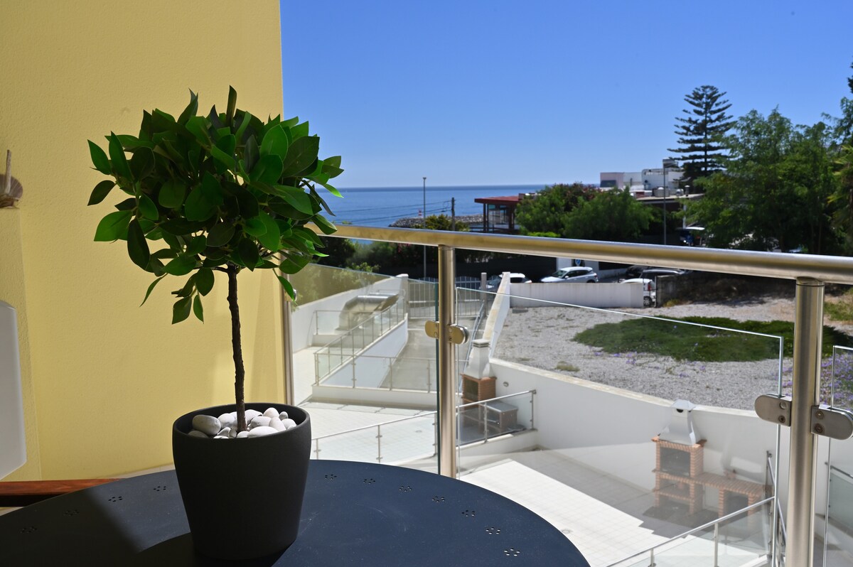 A potted plant with green leaves is positioned on a table, providing a natural touch. In the background, a clear view of the sea is visible, along with the surrounding greenery and residences. The space is flooded with natural light, enhancing the serene atmosphere.