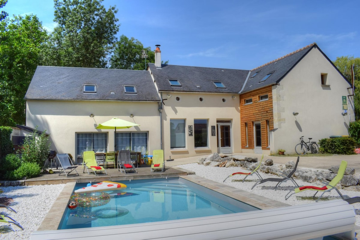 The exterior of a spacious gîte is highlighted, featuring a heated swimming pool surrounded by a sun terrace. Multiple lounge chairs are positioned around the pool, and a patio table with umbrellas offers shaded seating. Lush greenery and a bicycle are also visible beside the building.