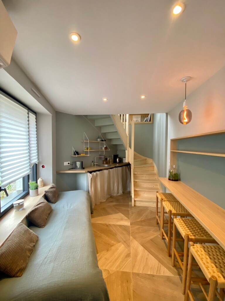 A compact living area features a light blue wall, modern lighting fixtures, and a small kitchenette at the far side. A staircase leads to the upper level, while a comfortable seating area with cushions is visible along the left. Two wooden bar stools are positioned at the counter.
