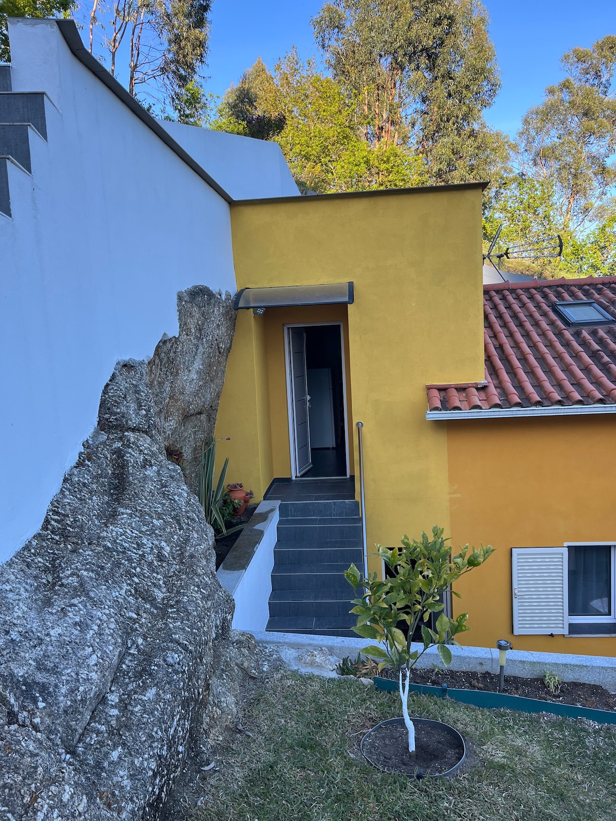 A pathway lined with soft grass leads up to a modern yellow building with a red-tiled roof. The entrance is framed by a large rock formation, complemented by greenery and trees in the surrounding area.
