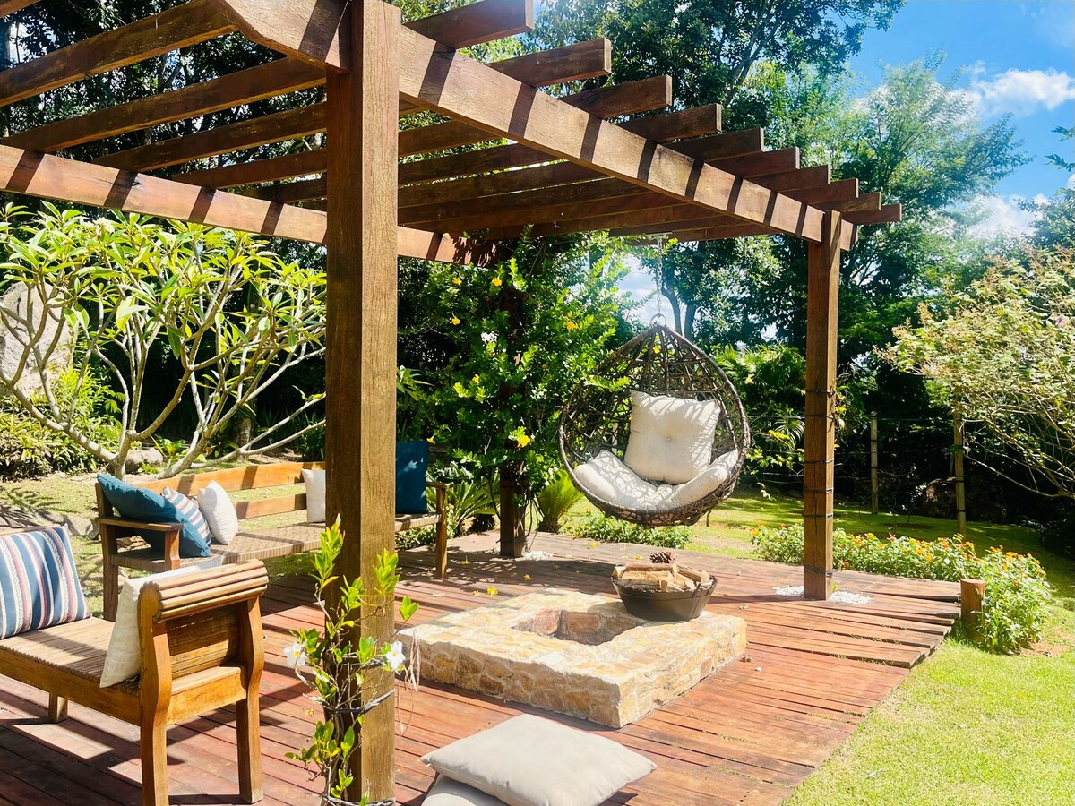 An outdoor area is showcased with a wooden pergola providing shade. A cozy swinging chair hangs from the structure, accompanied by seating options. A stone fire pit is situated on a wooden deck, surrounded by greenery and neatly arranged plants, encouraging relaxation in nature.