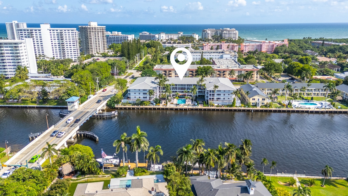 An aerial view showcases the coastal area, with the ocean visible in the background. The property overlooks a serene waterway, bordered by lush palm trees and well-maintained buildings. The community pool and surrounding amenities are evident, contributing to a relaxing atmosphere by the water.