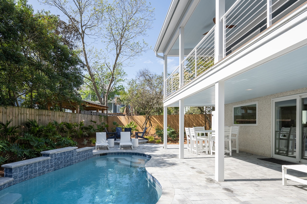 A private outdoor pool area features a sparkling swimming pool surrounded by paved stones. Lounge chairs are positioned nearby, and a covered dining area with a table is visible. Lush landscaping with trees and shrubs creates a serene environment, with the house's elevated balcony overlooking the space.