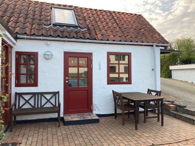 The exterior of a charming white guest annex is visible, featuring a red door and coordinated window frames. A wooden table and two chairs are placed on a paved terrace area, inviting outdoor relaxation. The sloped roof is adorned with red tiles, complemented by a skylight.
