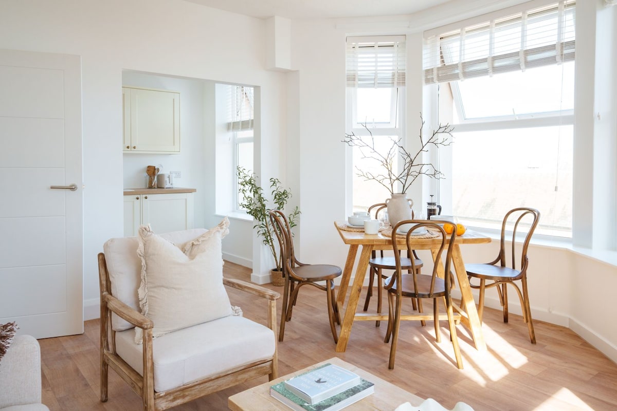 The open-plan living area features a bright dining space with a wooden table surrounded by four chairs. A cozy armchair is positioned nearby. Natural light streams through large windows adorned with elegant blinds, and greenery is displayed in a corner plant.
