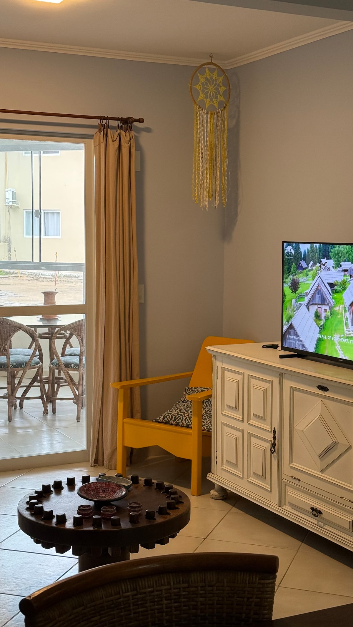 A living area is shown with light-colored walls and tiled flooring. A wooden coffee table with a circular design sits at the center, surrounded by woven chairs. Natural light enters through a sliding glass door leading to a patio, where outdoor seating is visible.
